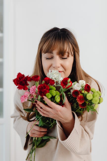 A woman with light brown hair and bangs, wearing a beige sweater, holds a vibrant bouquet of mixed fresh flowers close to her face with her eyes gently closed and a slight smile. The bouquet includes 