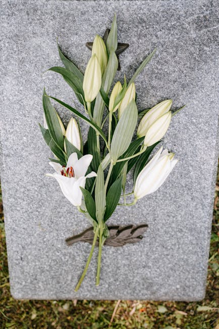 A floral tribute featuring white lilies and unopened lily buds arranged on a grey stone memorial plaque. The white lilies have broad petals with some fully open, revealing deep red stamens, while othe