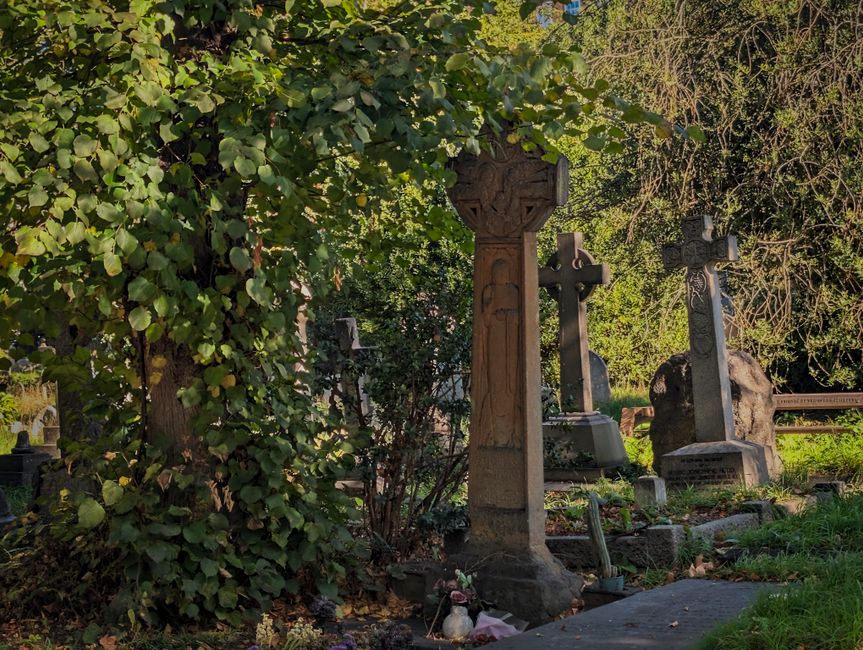 A floral tribute arrangement featuring a variety of fresh flowers in soft pastel colors, including pink and cream roses, lavender, and greenery, is placed on a gravel path amid a historic cemetery sce