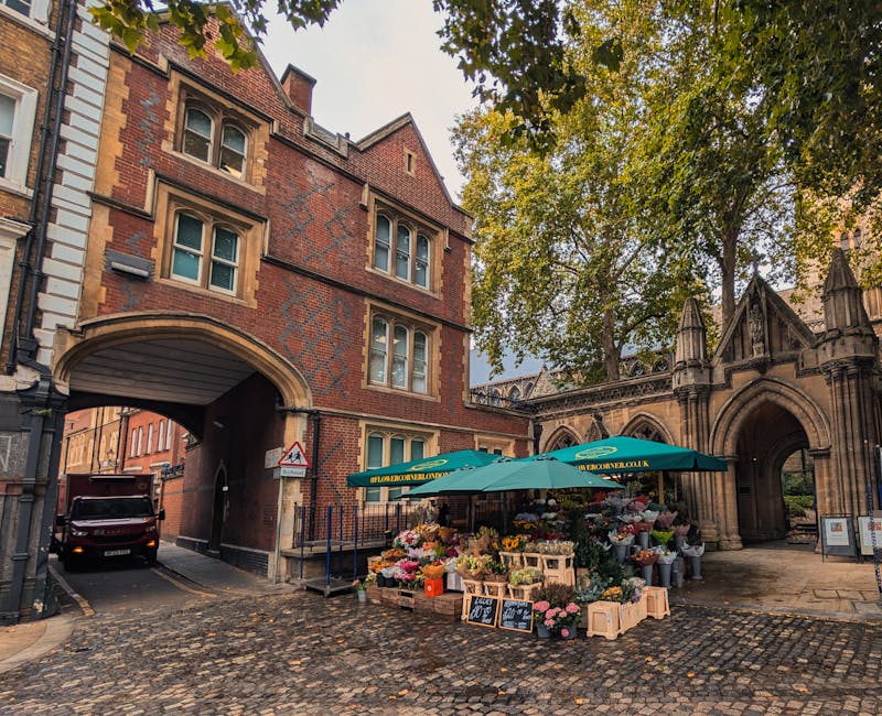 An outdoor flower stall set up on a cobblestone street near a historic stone archway, offering a vibrant selection of fresh floral arrangements including roses, peonies, lilies, and sunflowers in vari